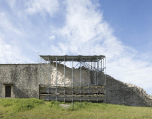 Struttura metallica di sostegno su muro parzialmente crollato in pietra con prato e cielo sereno.